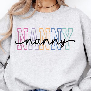 May include: A light gray sweatshirt with the word "NANNY" in colorful block letters and a cursive "nanny" underneath. The letters are outlined in pink, blue, orange, and green. The sweatshirt has a crew neck.