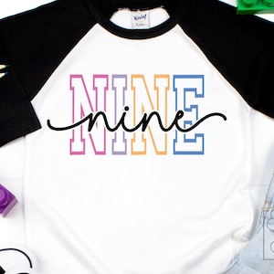 May include: White and black raglan style t-shirt with the word "NINE" in colorful block letters and a cursive "nine" underneath. The shirt is paired with blue jeans and colorful pencils.