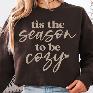 Puede incluir: Una sudadera marrón oscuro con la frase "tis the season to be cozy" en escritura beige claro. La sudadera tiene mangas largas y cuello redondo. El texto incluye un símbolo de corazón.