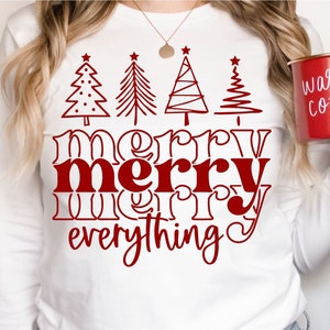 May include: White long-sleeve shirt with a red graphic design that says "merry merry everything" with stylized Christmas trees above the text. The shirt is being worn by a person holding a red enamel mug that says "warm cozy".