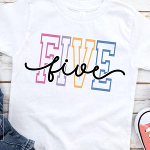 May include: White t-shirt with the word "FIVE" in colorful block letters and a cursive "five" underneath. The shirt is next to blue jeans and pink sneakers.