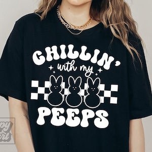 Könnte beinhalten: Schwarzes T-Shirt mit weißem Text, der "Chillin' with my peeps" in einer Retro-Schriftart lautet. Der Text ist von drei weißen Cartoon-Peeps und einem karierten Muster umgeben.
