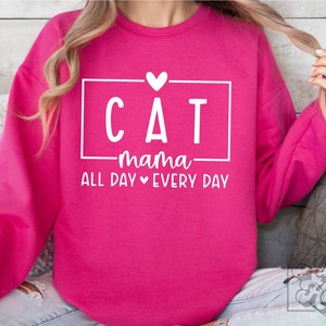 May include: A bright pink sweatshirt with the words "CAT mama ALL DAY EVERY DAY" in white, inside a white rectangle. A small white heart is above the word "mama". The sweatshirt is a crew neck style.