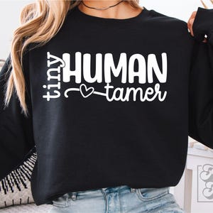 Può includere: Felpa nera con la scritta bianca "tiny HUMAN tamer" e un simbolo a forma di cuore. Uno stile di abbigliamento casual e confortevole. Il design è adatto all'uso quotidiano.
