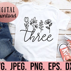 May include: White t-shirt with black floral designs and the word "three" in a cursive font.
