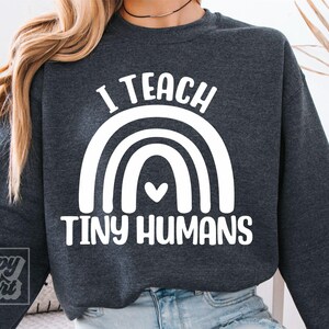 Può includere: Felpa grigio scuro con la scritta bianca "I TEACH TINY HUMANS" sopra un disegno di arcobaleno bianco con un cuore. La felpa è indossata da una persona con i capelli biondi.