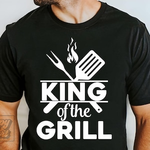 May include: Black t-shirt with a white graphic design featuring a fork, spatula, and flames above the words "KING of the GRILL". The design is centered on the chest.