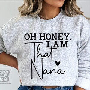K&ouml;nnte beinhalten: Grauer Pullover mit dem Text "Oh honey, I am that Nana" in Schwarz mit einem kleinen schwarzen Herz.
