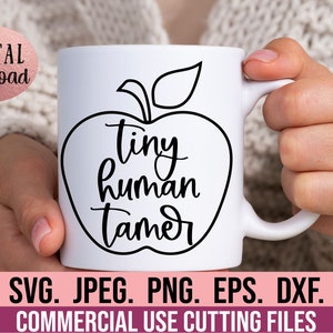 Può includere: Una tazza in ceramica bianca con un contorno nero di una mela e il testo "tiny human tamer" all'interno della mela. La tazza è tenuta da una mano.