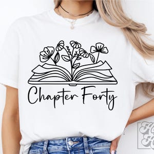 Può includere: Maglietta bianca con un contorno nero di un libro aperto con fiori che crescono da esso. Il testo "Chapter Forty" è sotto il libro.
