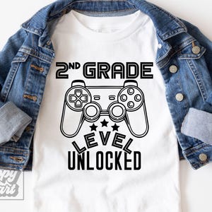 Puede incluir: Camiseta blanca con un gráfico negro de un mando de videojuegos y el texto "2nd Grade Level Unlocked". Una chaqueta vaquera azul enmarca la camiseta.