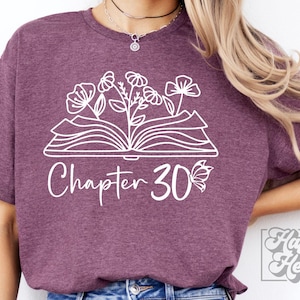 May include: A purple t-shirt with a white design of an open book with flowers growing out of it. The text "Chapter 30" is below the book with a butterfly to the right of the number.