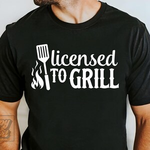 May include: Black t-shirt with the phrase "licensed TO GRILL" in white, accompanied by a spatula and flames graphic. The shirt is a casual wear item, perfect for grilling enthusiasts.
