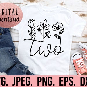 May include: A white t-shirt with a black design of the word "two" with stylized flowers. The shirt is on a wooden surface with a pair of pink sneakers.
