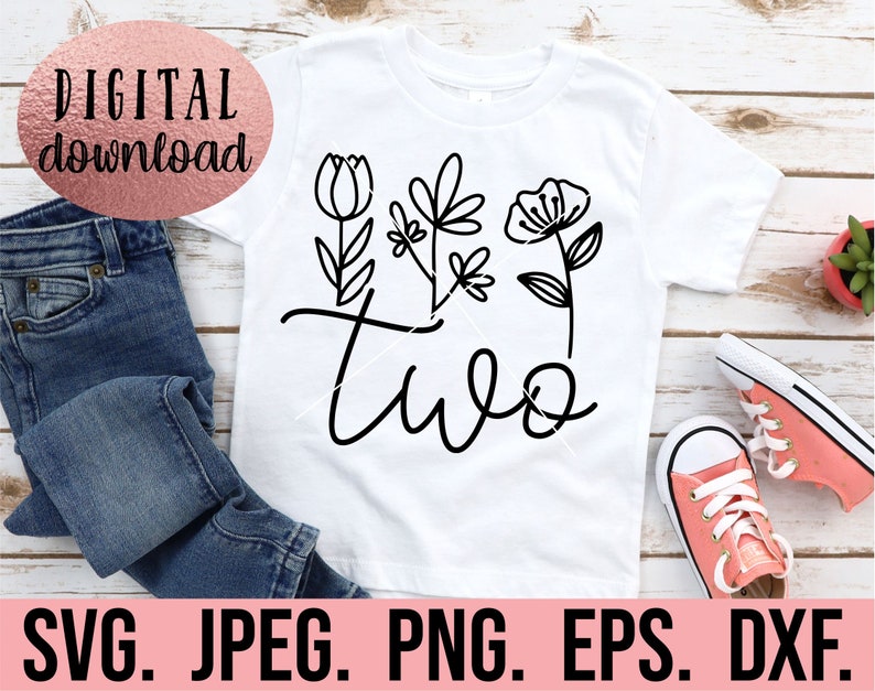 May include: A white t-shirt with a black design of the word "two" with stylized flowers. The shirt is on a wooden surface with a pair of pink sneakers.