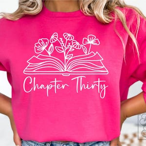 May include: A pink sweatshirt with a white design of an open book with flowers growing out of it. The text "Chapter Thirty" is below the book.
