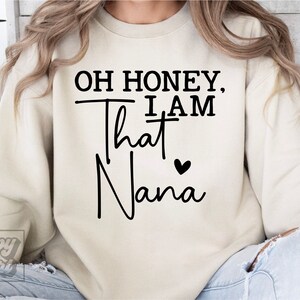 K&ouml;nnte beinhalten: Ein cremefarbener Pullover mit dem Text "Oh honey, I am that Nana" in schwarzer Schrift mit einem kleinen schwarzen Herz.