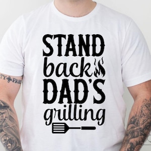 May include: White t-shirt with the text "STAND back DAD'S grilling" in bold black letters. The design includes flames and a spatula graphic, ideal for a barbecue enthusiast.