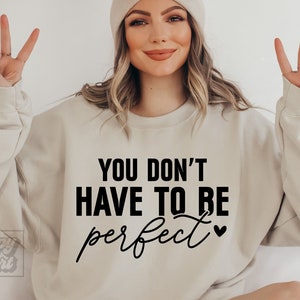 Könnte beinhalten: Ein beiger Pullover mit dem Text "YOU DON'T HAVE TO BE perfect" in schwarzer Schrift mit einem kleinen Herzsymbol.