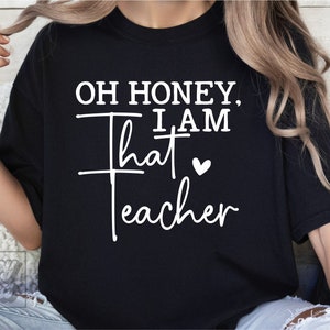 May include: Black t-shirt with white text that reads "Oh honey, I am that teacher" with a small white heart.