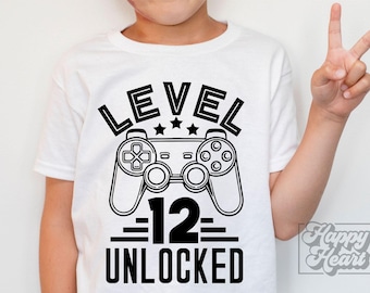 Könnte beinhalten: Weißes T-Shirt mit einem schwarzen Grafikdesign mit den Worten "LEVEL 12 UNLOCKED" und einem Controller-Symbol. Das Design ist auf dem T-Shirt zentriert. Eine Hand macht das Friedenszeichen.