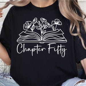 Puede incluir: Camiseta negra con un diseño blanco de un libro abierto con flores que crecen de él. El texto "Chapter Fifty" está escrito debajo del libro.