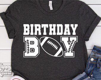 May include: A dark gray t-shirt with the text "BIRTHDAY BOY" in white. The "B" and "Y" are stylized with stitching details. A white football is centered between the letters.