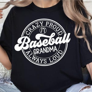 May include: Black t-shirt with a white circular graphic that reads "Crazy Proud Baseball Grandma Always Loud" with a baseball illustration. The shirt is a casual style.