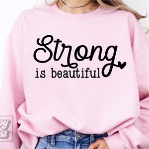 May include: A light pink sweatshirt with the words "Strong is beautiful" in black script. The sweatshirt has a crew neck and long sleeves. The brand logo "Happy Heart" is in the bottom left corner.