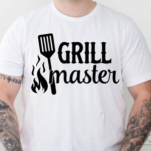 Puede incluir: Camiseta blanca con un gráfico negro que dice "GRILL master". El diseño incluye una espátula y llamas. La camiseta es de estilo casual.