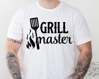 May include: White t-shirt with a black graphic that reads "GRILL master". The design includes a spatula and flames. The shirt is a casual wear item.
