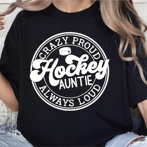 May include: Black t-shirt with a white circular graphic that reads "CRAZY PROUD Hockey AUNTIE ALWAYS LOUD". The design includes a hockey puck illustration. The shirt is a casual wear item.