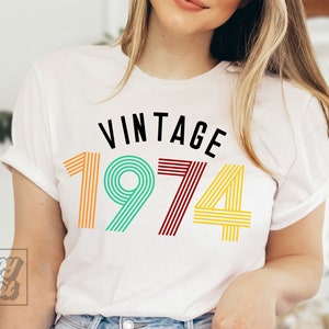 May include: White t-shirt with the text "Vintage 1974" in black and colorful striped font.