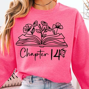 Puede incluir: Una sudadera rosa brillante con un gráfico negro de un libro abierto con flores y el texto "Chapter 14". La sudadera es de color liso y tiene mangas largas.