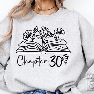 May include: A gray sweatshirt with a black and white design of an open book with flowers growing out of it. The text "Chapter 30" is below the book with a butterfly to the right of the number.