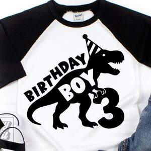 Könnte beinhalten: Weißes und schwarzes Raglan-Shirt mit einem schwarzen Dinosaurier-Motiv, das einen Partyhut trägt. Der Text "BIRTHDAY BOY 3" ist auf dem Shirt aufgedruckt. Eine blaue Jeans und schwarze Turnschuhe sind ebenfalls zu sehen.