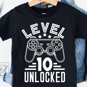 Puede incluir: Camiseta negra con un gráfico blanco que presenta un mando de videojuegos, las palabras "LEVEL 10 UNLOCKED" y tres estrellas. La camiseta está colgada en una percha de madera.