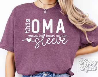 May include: A maroon t-shirt with the text "this OMA wears her heart on her sleeve" in white script. The shirt has a relaxed fit and short sleeves. The design includes a heart symbol.