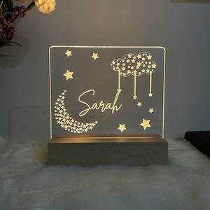 Könnte beinhalten: Ein weißes Acryl-Nachtlicht mit einem Halbmond, Sternen und einem Wolkenmuster. Der Name "Sarah" ist in das Licht graviert. Das Licht steht auf einem beigefarbenen Sockel.