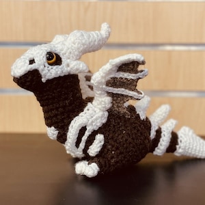 May include: A crocheted dragon toy with a brown body and white skeletal details. The dragon has a white horn and a white tail.