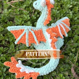 May include: A crocheted dragon plush toy featuring a light mint green body with bright orange accents along the spine, wings, and tail, and white detailing. The text "The Archaic Dragon - madisondinomay 2026" and "PATTERN!" are visible on the image.