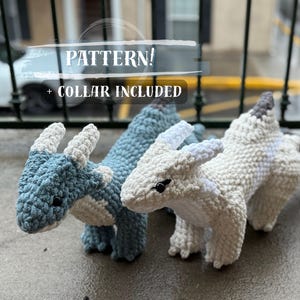 Puede incluir: Dos juguetes de dragón de ganchillo, uno azul claro y otro blanco. Ambos tienen cuernos y detalles blancos. El texto "PATTERN! + COLLAR INCLUDED" es visible. Los dragones están sobre una superficie de hormigón.