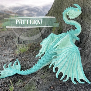 May include: A crocheted dragon amigurumi toy with a light blue and green color scheme. The dragon has a long neck, large wings, and a tail with spikes. The toy is sitting on a tree trunk with the word "PATTERN!" written on a white banner.