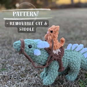 May include: A crocheted green dinosaur toy with a removable orange cat and saddle. The dinosaur features white accents and a brown bridle. The image includes the text "PATTERN! + REMOVABLE CAT & SADDLE".