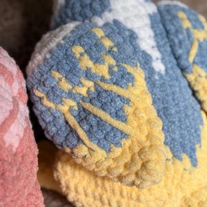 May include: Close-up of a plush, crocheted bird with blue, yellow, and white details. The wings are a mix of blue and yellow, with white accents. The word "PATTERN!" is visible in the top left corner.