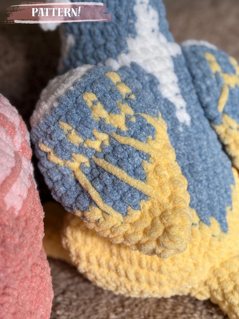May include: Close-up of a plush, crocheted bird with blue, yellow, and white details. The wings are a mix of blue and yellow, with white accents. The word "PATTERN!" is visible in the top left corner.