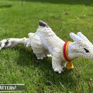 May include: A white crocheted dragon toy with a gray and black tail, lying on green grass. The dragon has a red and yellow collar with a small gold bell. The word "PATTERN!" is at the bottom of the image.