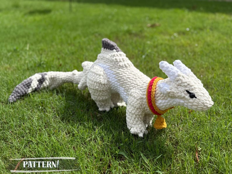 May include: A white crocheted dragon toy with a gray and black tail, lying on green grass. The dragon has a red and yellow collar with a small gold bell. The word "PATTERN!" is at the bottom of the image.