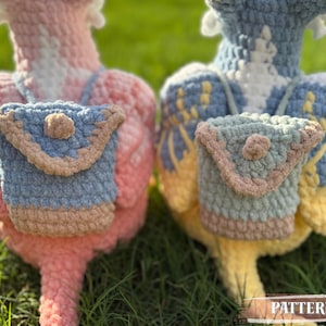 May include: Two handmade crochet bird figures, one pink and one blue, each with a small matching backpack. The backpacks are light blue with tan trim and a button closure. The birds are on a grassy surface. The word "PATTERN!" is visible.