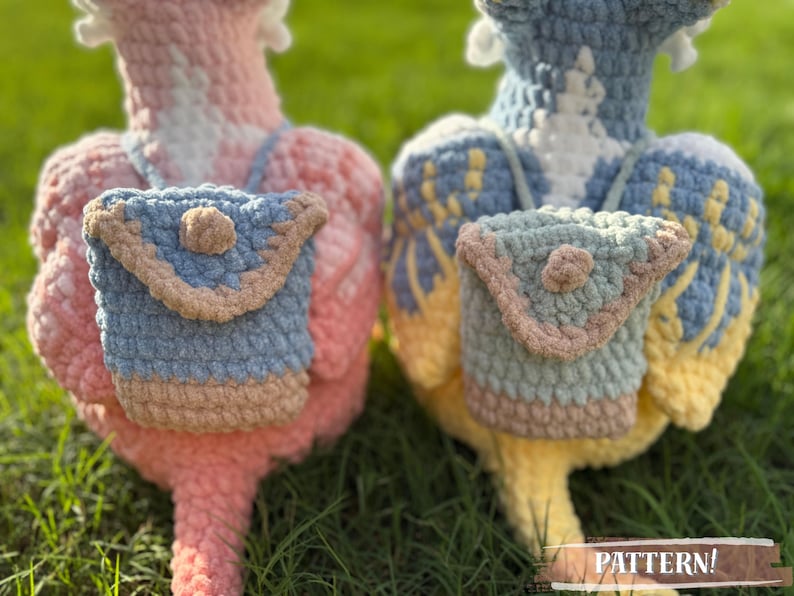 May include: Two handmade crochet bird figures, one pink and one blue, each with a small matching backpack. The backpacks are light blue with tan trim and a button closure. The birds are on a grassy surface. The word "PATTERN!" is visible.